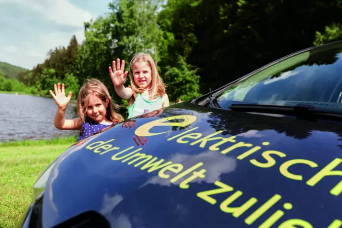 Das Bild zeigt winkende Kinder hinter der Motorhaube eines E-Autos