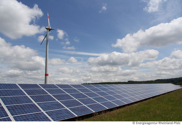 Im Vordergrund steht eine Freiflächen-Photovoltaik-Anlage und im Hintergrund ein Windrad