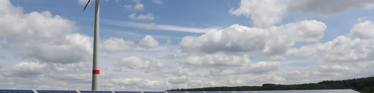 Im Vordergrund steht eine Freiflächen-Photovoltaik-Anlage und im Hintergrund ein Windrad