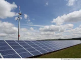 Im Vordergrund steht eine Freiflächen-Photovoltaik-Anlage und im Hintergrund ein Windrad