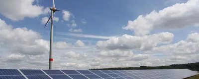 Im Vordergrund steht eine Freiflächen-Photovoltaik-Anlage und im Hintergrund ein Windrad