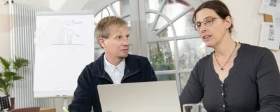 Zwei Personen sitzen an einem Tisch und blicken auf einen Laptop. Dabei unterhalten sie sich.