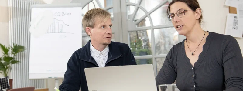 Zwei Personen sitzen an einem Tisch und blicken auf einen Laptop. Dabei unterhalten sie sich.