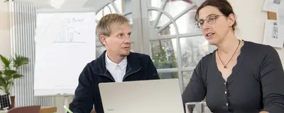 Zwei Personen sitzen an einem Tisch und blicken auf einen Laptop. Dabei unterhalten sie sich.