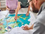 Mehrere Menschen sitzen an einem Tisch. Auf dem Tisch liegt ein großer Plan. Darauf steht ein Windrand. Jemand deutet mit dem Finger auf den Plan.