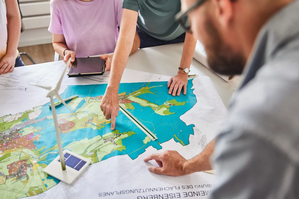 Mehrere Menschen sitzen an einem Tisch. Auf dem Tisch liegt ein großer Plan. Darauf steht ein Windrand. Jemand deutet mit dem Finger auf den Plan.