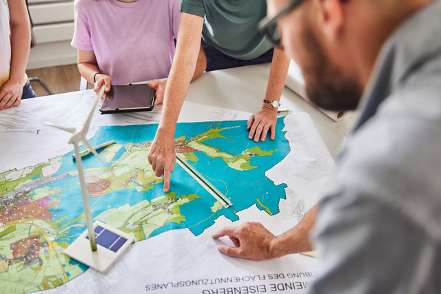 Mehrere Menschen sitzen an einem Tisch. Auf dem Tisch liegt ein großer Plan. Darauf steht ein Windrand. Jemand deutet mit dem Finger auf den Plan.