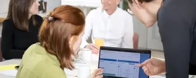 Mehrere Personen sitzen an einem Tisch. Zwei Personen deuten auf einen Laptop und besprechen etwas.
