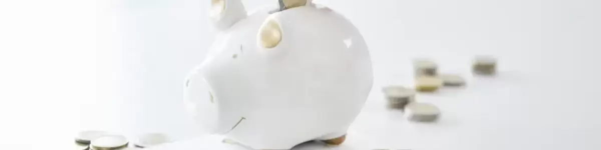 Eine Hand wirft eine Münze in ein Sparschwein. Auf dem Tisch liegen weitere Münzen, ein Notizblock, ein Taschenrechner und ein Kugelschreiber