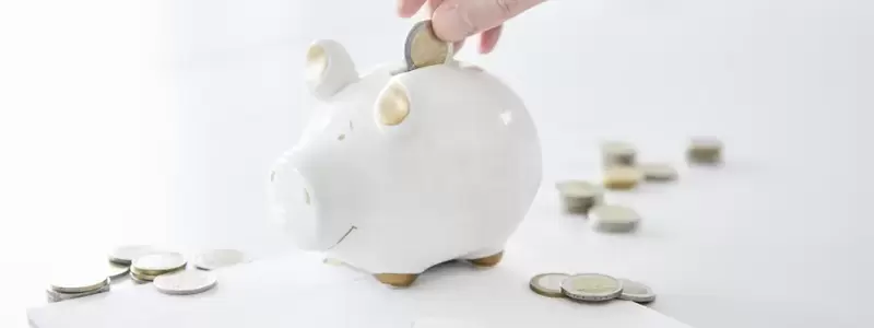 Eine Hand wirft eine Münze in ein Sparschwein. Auf dem Tisch liegen weitere Münzen, ein Notizblock, ein Taschenrechner und ein Kugelschreiber