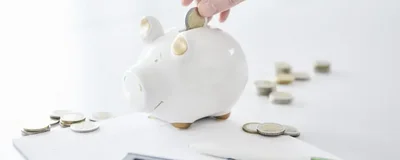 Eine Hand wirft eine Münze in ein Sparschwein. Auf dem Tisch liegen weitere Münzen, ein Notizblock, ein Taschenrechner und ein Kugelschreiber