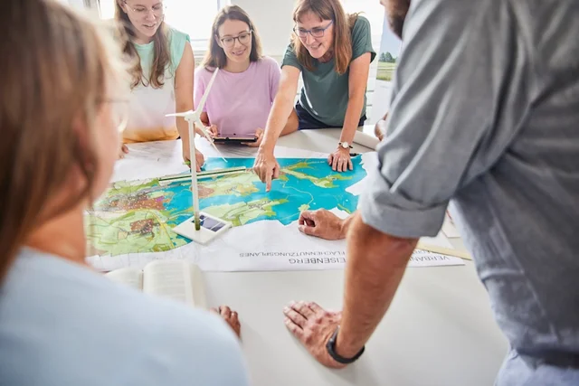 Mehrere Menschen sind um einen Tisch versammelt und sehen auf einen Flächennutzungsplan. Darauf steht auch das Modell eines Windrades. 