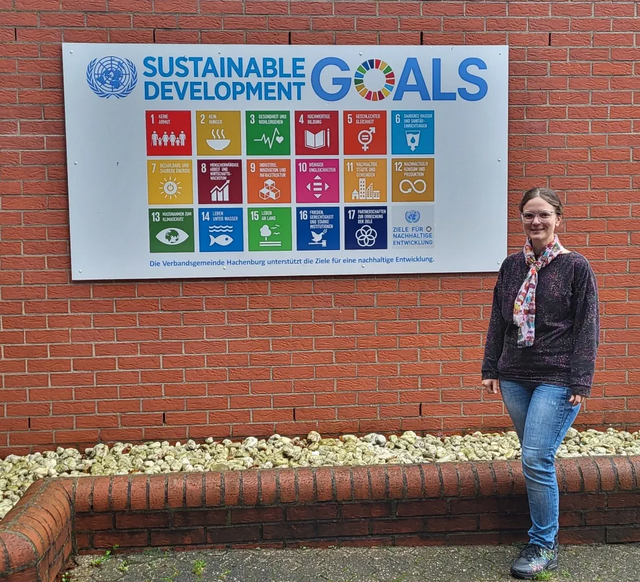 Frau steht vor einer Hauswand, an der eine Tafel mit Nachhaltigkeitszielen hängt