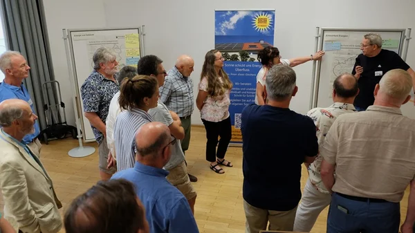 Mehrere Menschen stehen in einem Raum und hören einer Frau zu, die anhand eines Flipcharts etwas erklärt