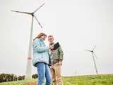 Zwei Menschen stehen auf einem Feld. Im Hintergrund stehen zwei Windräder. Die beiden Menschen blicken auf ein tablet und besprechen sich.