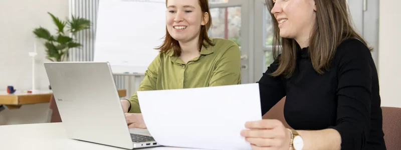 Zwei Menschen sitzen am Tisch und unterhalten sich. Eine Person hat einen Laptop, die andere ein Blatt Papier in der Hand
