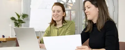 Zwei Menschen sitzen am Tisch und unterhalten sich. Eine Person hat einen Laptop, die andere ein Blatt Papier in der Hand