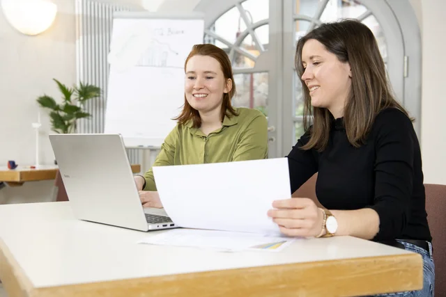 Zwei Menschen sitzen am Tisch und unterhalten sich. Eine Person hat einen Laptop, die andere ein Blatt Papier in der Hand