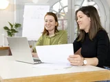 Zwei Menschen sitzen am Tisch und unterhalten sich. Eine Person hat einen Laptop, die andere ein Blatt Papier in der Hand