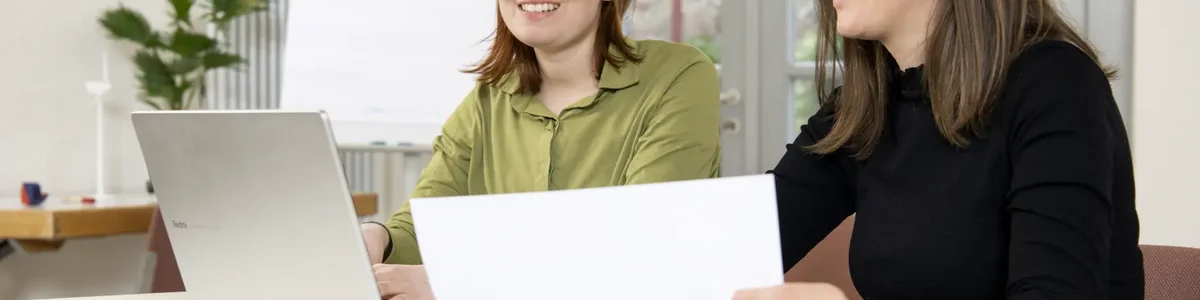 Zwei Menschen sitzen am Tisch und unterhalten sich. Eine Person hat einen Laptop, die andere ein Blatt Papier in der Hand