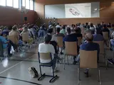 Viele Menschen sitzen auf Stühlen in einer Halle und hören einem Vortragenden zu