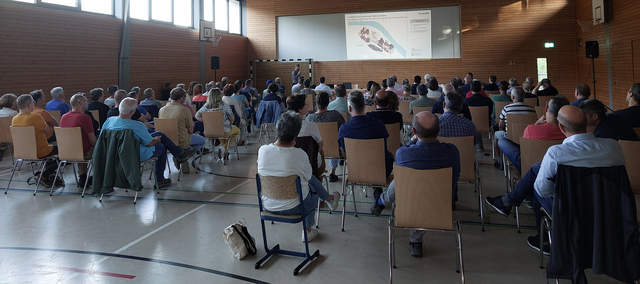 Viele Menschen sitzen auf Stühlen in einer Halle und hören einem Vortragenden zu