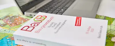 Das Baugesetzbuch liegt auf einem Tisch, daneben ein Bauplan und ein Laptop.
