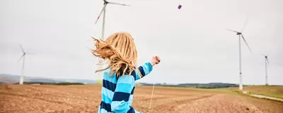 ein Kind lässt auf einem Feld einen Drachen steigen, im Hintergrund sind Windräder zu sehen