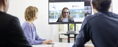 Drei Menschen sitzen in einer Besprechung. Eine Person ist virtuell zugeschaltet, das Gesicht ist auf einem Bildschirm.