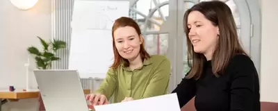 Zwei Menschen sitzen an einem Tisch und besprechen etwas. Eine Person sitzt vor dem Laptop und tippt, die andere zeigt auf ein Blatt Papier.