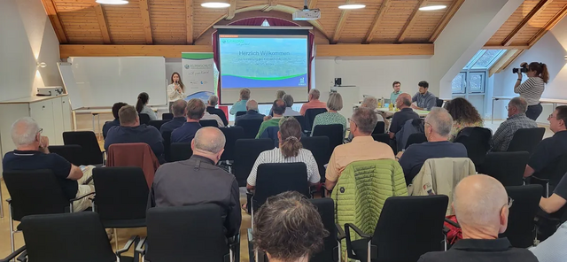 Viele Menschen sitzen in einem Raum und schauen sich eine Präsentation auf einem großen Bildschirm an