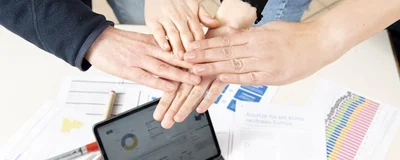Auf einem Tisch steht ein Laptop und verschiedene Arbeitsmaterialien. Darüber treffen sich die Hände von vier Menschen in der Mitte.