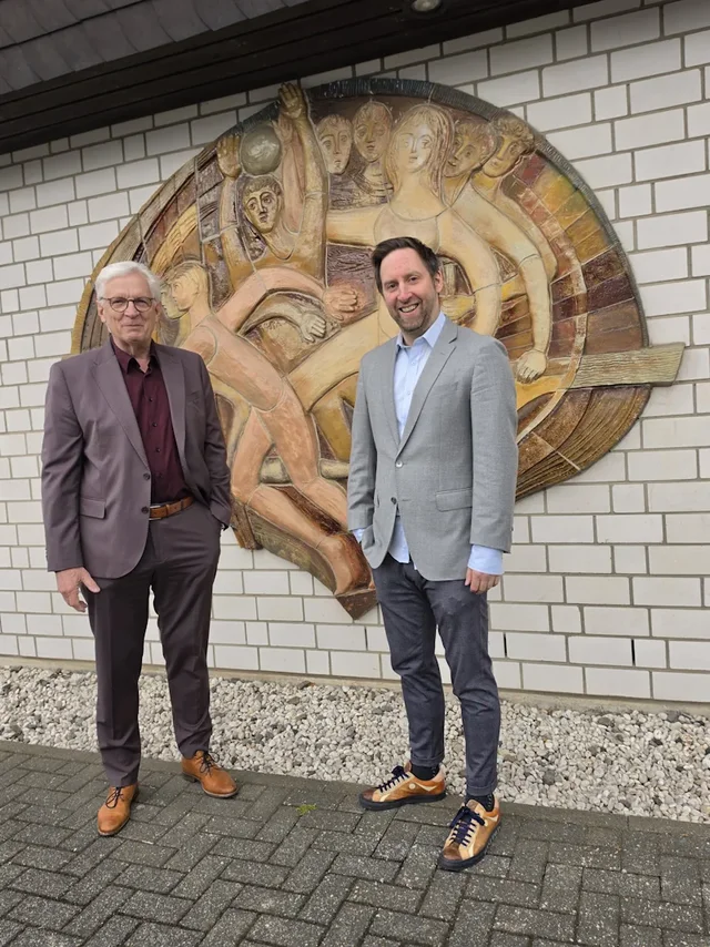 Zwei Männer stehen vor einer Mauer und schauen in die Kamera