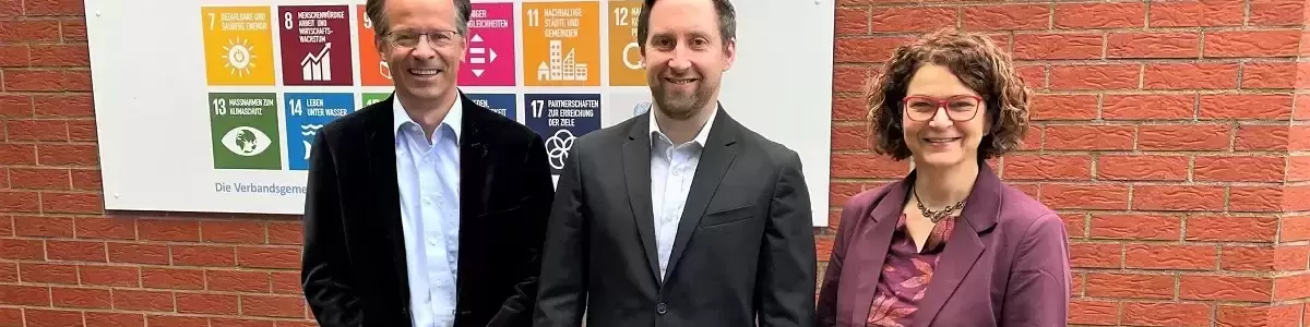 r Erste Beigeordnete Marco Dörner, Nachhaltigkeitskoordinator Timo Karl und die Bürgermeisterin der Verbandsgemeinde Hachenburg, Gabriele Greis, vor der Tafel mit den globalen Nachhaltigkeitszielen, die gut sichtbar am Eingang des Verwaltungsgebäudes angebracht worden sind.  