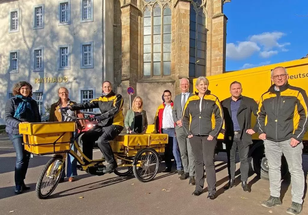 Beschäftigte der Post auf E-Bikes, der Stadtverwaltung und andere Personen stehen draußen vor einem Streetscooter