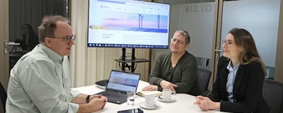 Drei Menschen sitzen in einem Besprechungsraum. Vor einer Person steht ein Laptop, im Hintergrund ist das Bild des Laptops auf einen Bildschirm gespiegelt. Die Person mit dem Laptop spricht zu den anderen beiden Personen und berät sie.