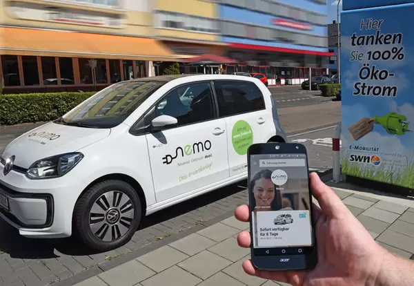 Carsharing-Pkw im Hintergrund. Im Vordergrund eine Hand mit Smartphone und Carsharing-App