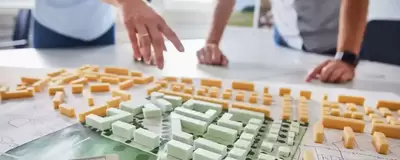 Das Modell einer Stadt liegt auf dem Tisch. Eine Person, die am Tisch steht, zeigt auf das Modell. Eine weitere Person hat die Hände am Tisch aufgestützt.