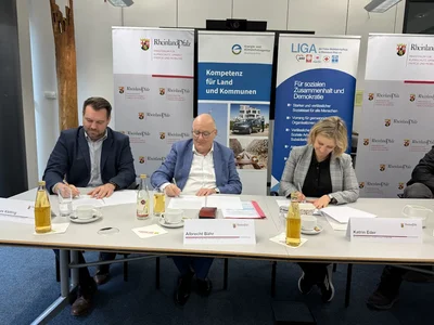 Zwei Männer und eine Frau sitzen an einem Tisch und unterschreiben alle ein Dokument