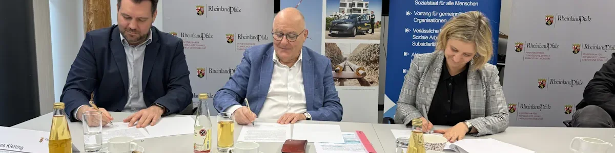 Zwei Männer und eine Frau sitzen an einem Tisch und unterschreiben alle ein Dokument