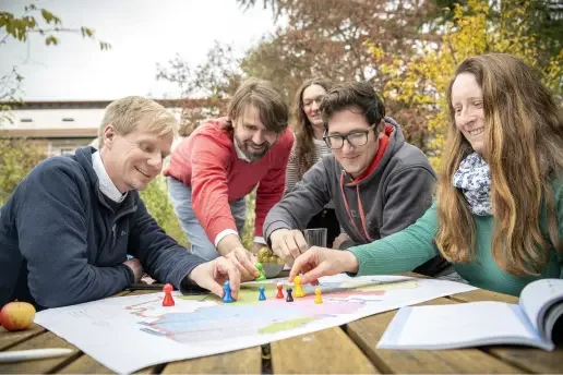 Auf einem Tisch liegt eine Karte von Rheinland-Pfalz. Fünf Menschen sitzen um den Tisch und platzieren auf der Karte bunte Kegel in menschenähnlicher Form.