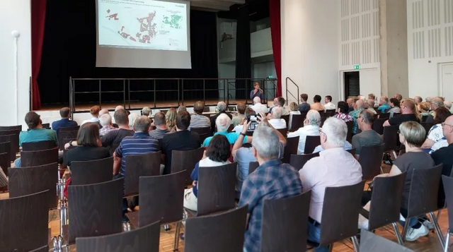 Viele Menschen sitzen in einem Raum und sehen sich eine Bildschirm-Präsentation an