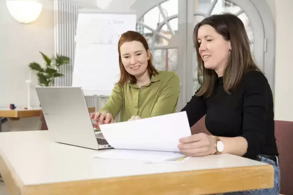 Zwei Menschen sitzen an einem Tisch und besprechen etwas. Eine Person sitzt vor dem Laptop und tippt, die andere zeigt auf ein Blatt Papier.