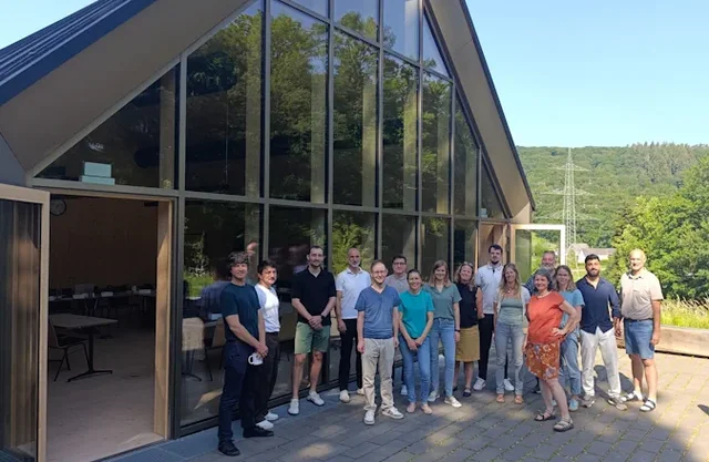Mehrere Menschen stehen beisammen vor einem Haus und schauen in die Kamera