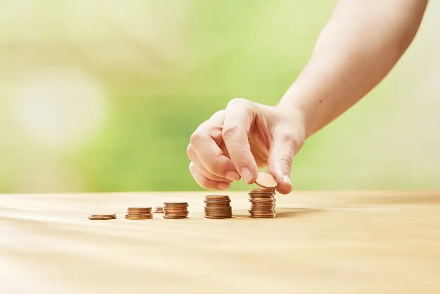 Auf einem Tisch stehen fünf unterschiedlich hohe Stapel mit cent-Münzen. Eine Hand legt auf den letzten und höchsten Stapel eine weitere Münze.