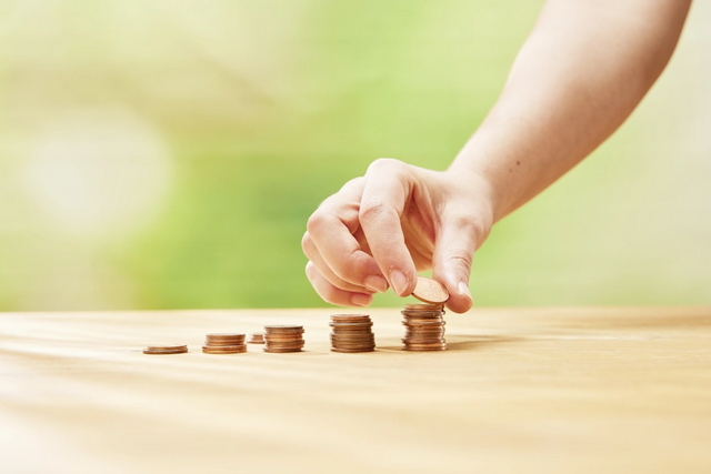 Auf einem Tisch stehen fünf unterschiedlich hohe Stapel mit cent-Münzen. Eine Hand legt auf den letzten und höchsten Stapel eine weitere Münze.