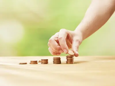 Auf einem Tisch stehen fünf unterschiedlich hohe Stapel mit cent-Münzen. Eine Hand legt auf den letzten und höchsten Stapel eine weitere Münze.