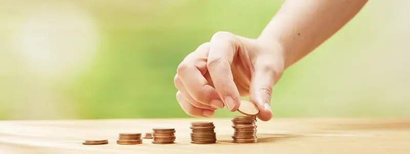 Auf einem Tisch stehen fünf unterschiedlich hohe Stapel mit cent-Münzen. Eine Hand legt auf den letzten und höchsten Stapel eine weitere Münze.