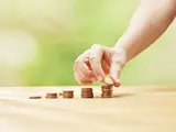 Auf einem Tisch stehen fünf unterschiedlich hohe Stapel mit cent-Münzen. Eine Hand legt auf den letzten und höchsten Stapel eine weitere Münze.