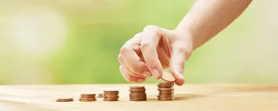 Auf einem Tisch stehen fünf unterschiedlich hohe Stapel mit cent-Münzen. Eine Hand legt auf den letzten und höchsten Stapel eine weitere Münze.
